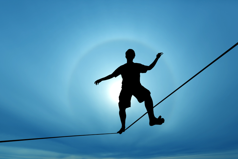 Silhouette of a young man walking on a tight rope with blue sky behind him.